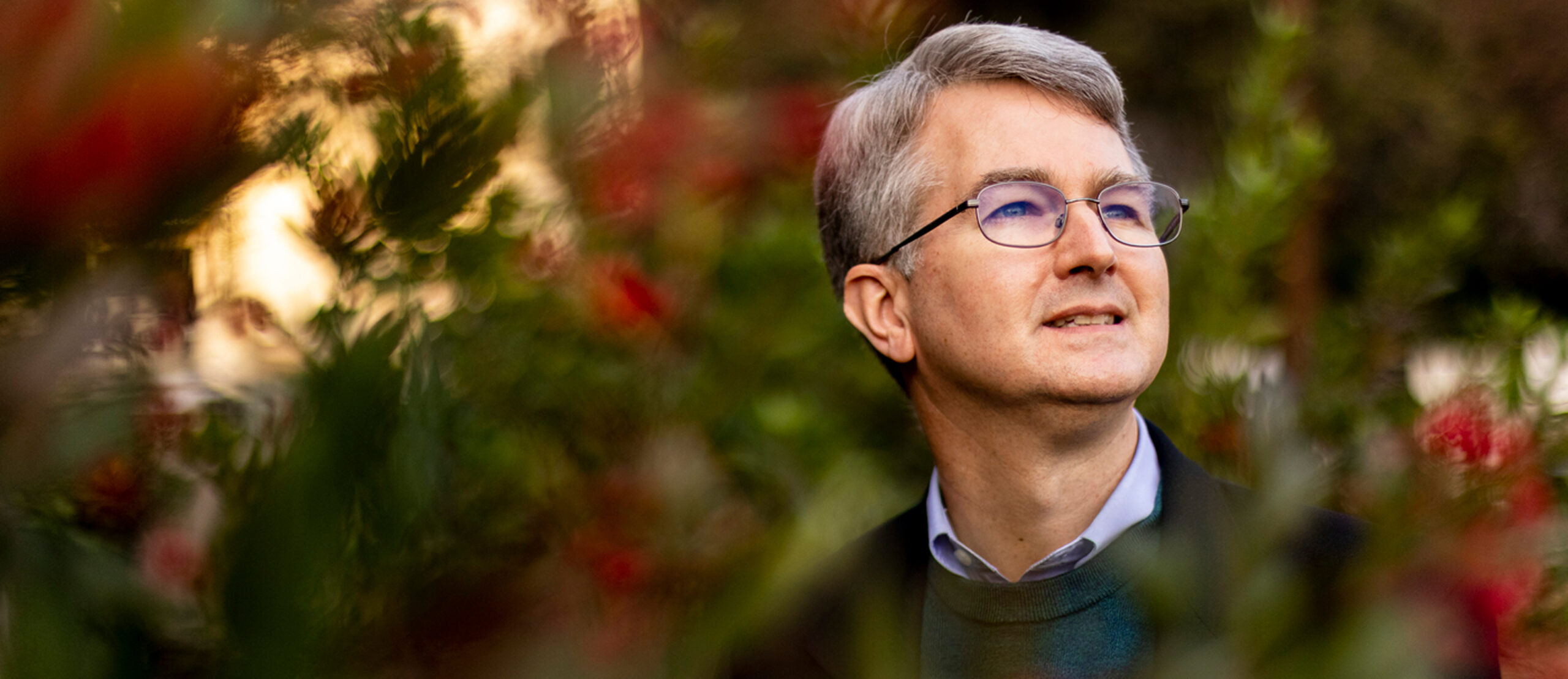 Mark Henderson, looks to the side while standing outdoors among green foliage and red flowers, with soft sunlight in the background.
