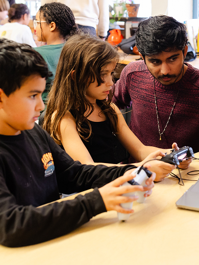 A group of students and a teacher gather around a laptop at a classroom table. Two children hold controllers, focused on the screen, while two adults assist and observe. Other students work in the background.