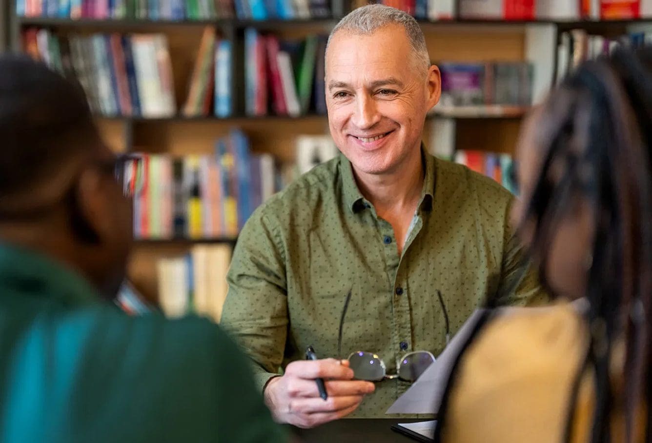 A smiling man in a green shirt sits at a table holding eyeglasses and a pen, speaking with two people in a library or study setting, with bookshelves in the background.