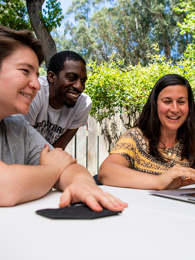 Three people sit and smile around a laptop outdoors at a table, with trees and sunlight in the background. They appear engaged and happy, looking at the computer screen together.