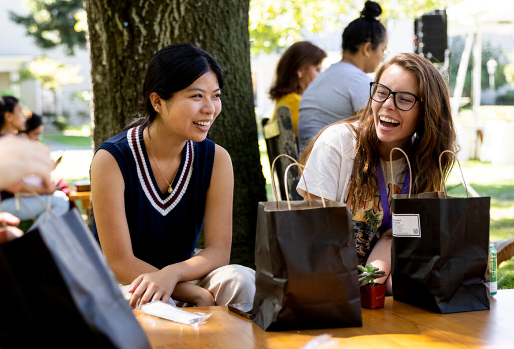 Two young women sit at an outdoor table, smiling and laughing together. Black lunch bags and a small potted plant are on the table. Sunlight filters through trees, and other people are visible in the background.