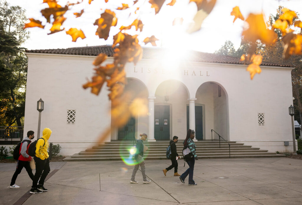 Students walk past a white building with arched doorways, labeled "LISSER HALL," as sunlight shines through autumn leaves in the foreground.