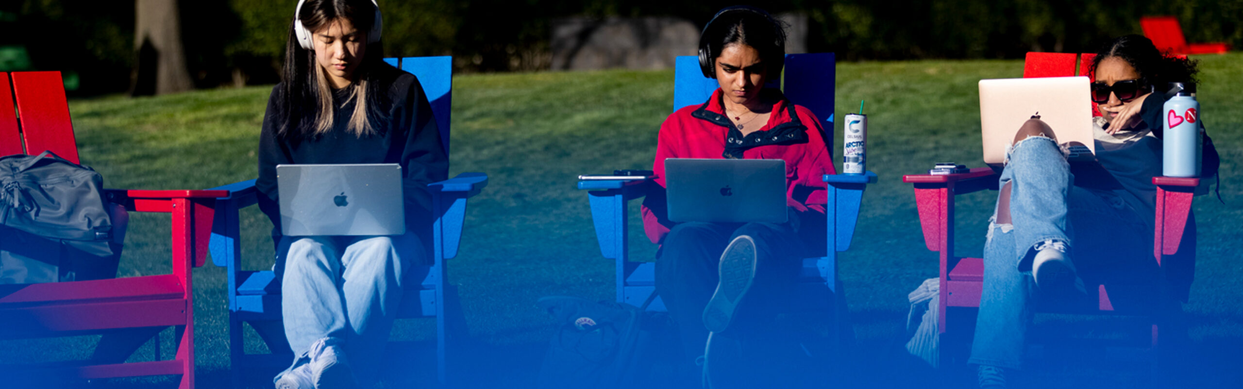 Three students sit outdoors on red and blue chairs, using laptops. One wears headphones, another has a drink can nearby, and one has sunglasses and a large water bottle. Grass and trees are visible in the background.