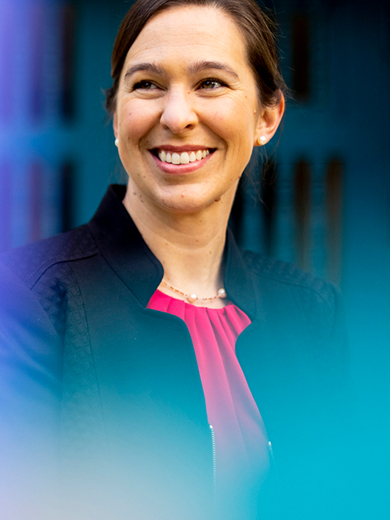 Dean Beth Kochly, wearing a dark jacket, smiles while looking to her left. The background is blurred with a blue door and a soft violet gradient on the left side of the image.