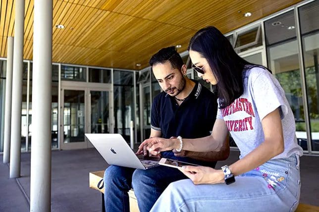 Two people sit on a bench outside the Graduate School of Business on Northeastern's Oakland campus, looking at a laptop together. One person points at the laptop screen while the other watches attentively. Both appear focused and engaged in their task.
