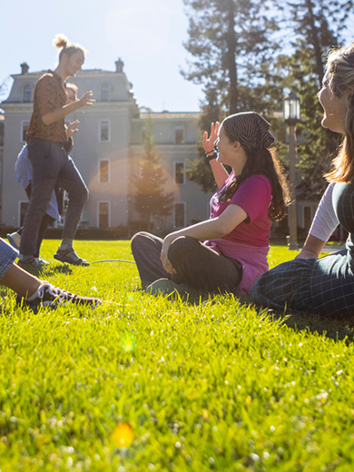 Four people sit and chat on a sunny lawn in front of Mills Hall, with a small dog nearby. Two others stand in the background. Trees and bright sunlight complete the scene.