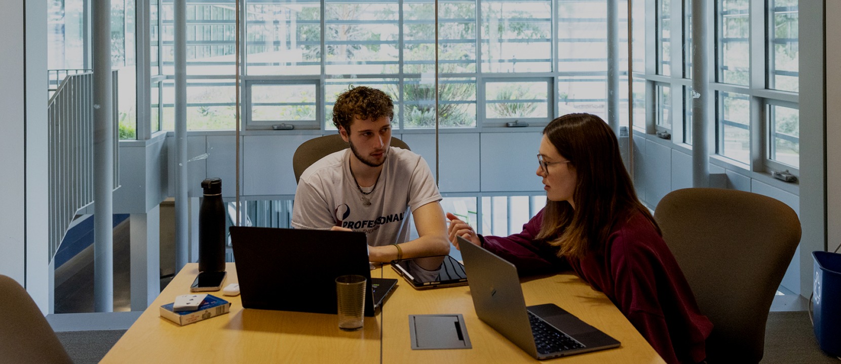 Two people sit across from each other at a table in a modern office, both working on laptops. They appear to be having a discussion. A water bottle, glass, and some items are also on the table.