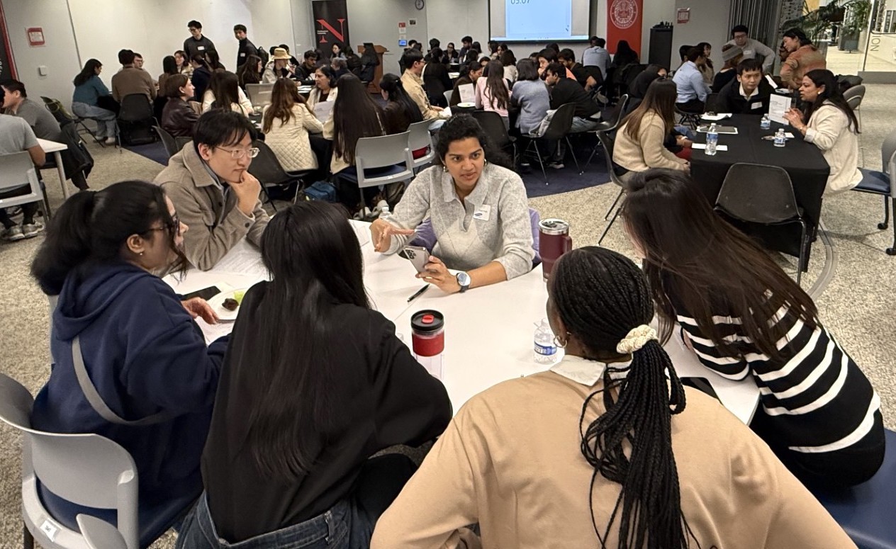A large group of people sit in small groups around tables at a graduate speed mentoring event. Some are taking notes, while others listen attentively. A screen and banners are visible in the background.