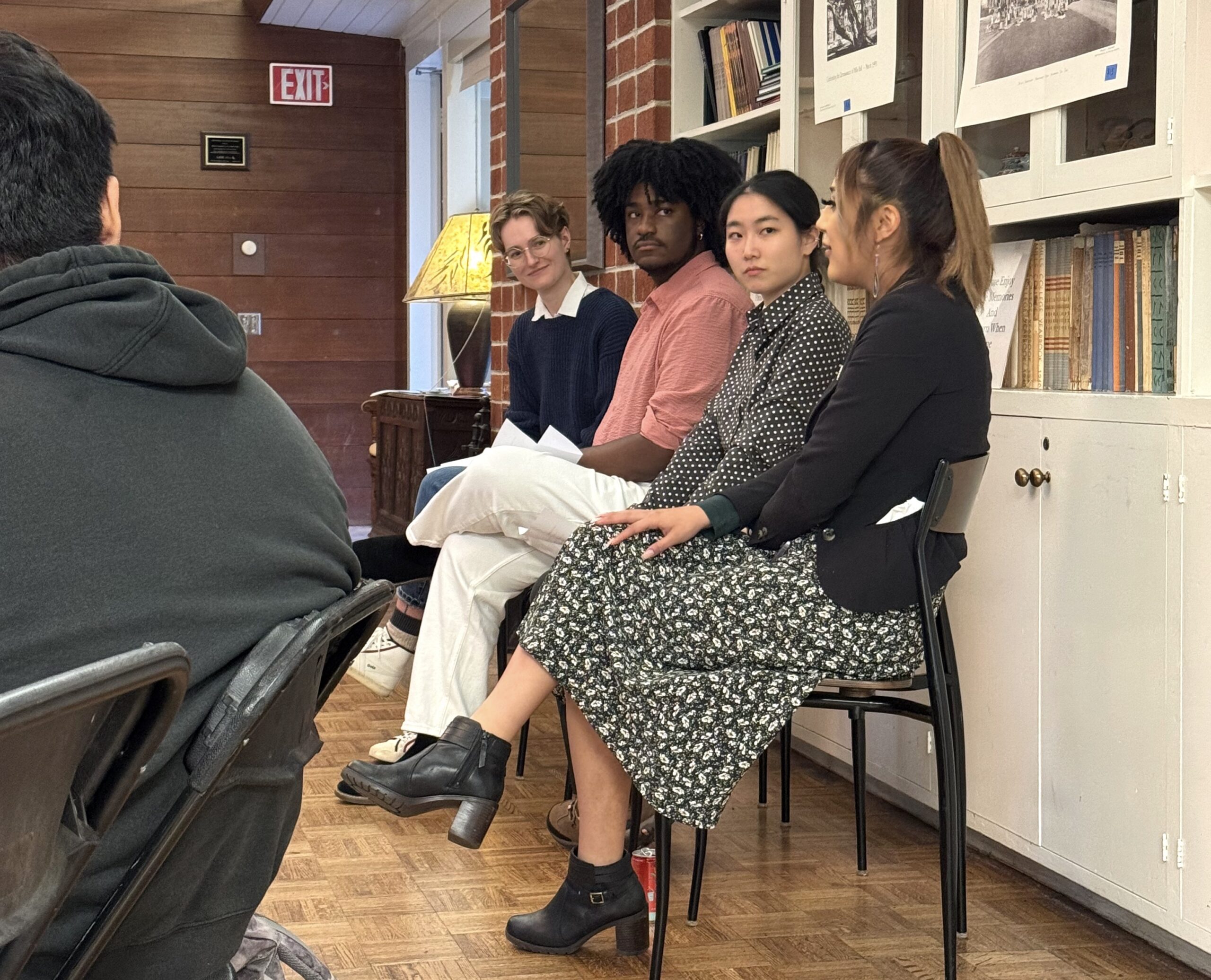 Four graduate students sit side by side on chairs in front of a group, participating in a panel at the Reinhardt Alumnae House on Northeastern University's Oakland campus. Bookshelves and wooden walls, with a lamp and framed photos are behind them.