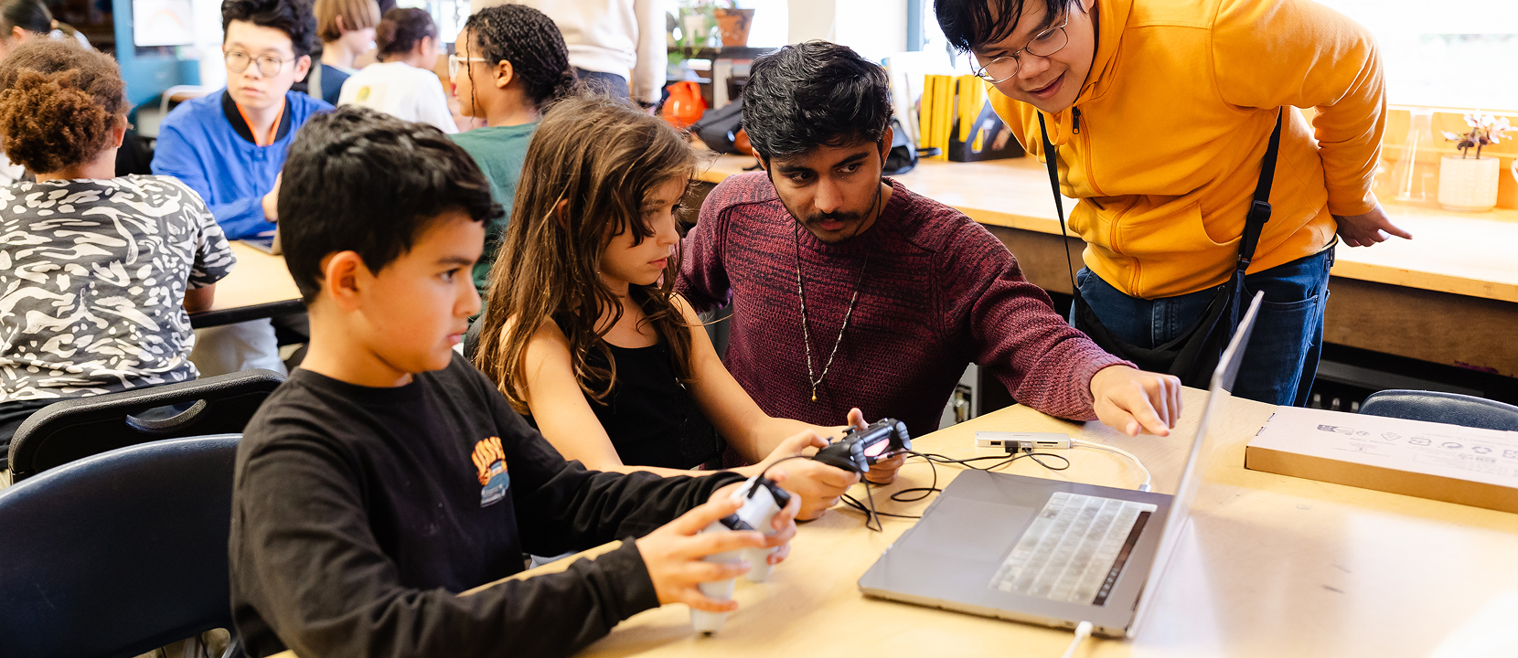 A group of students and a teacher gather around a laptop at a classroom table. Two children hold controllers, focused on the screen, while two adults assist and observe. Other students work in the background.