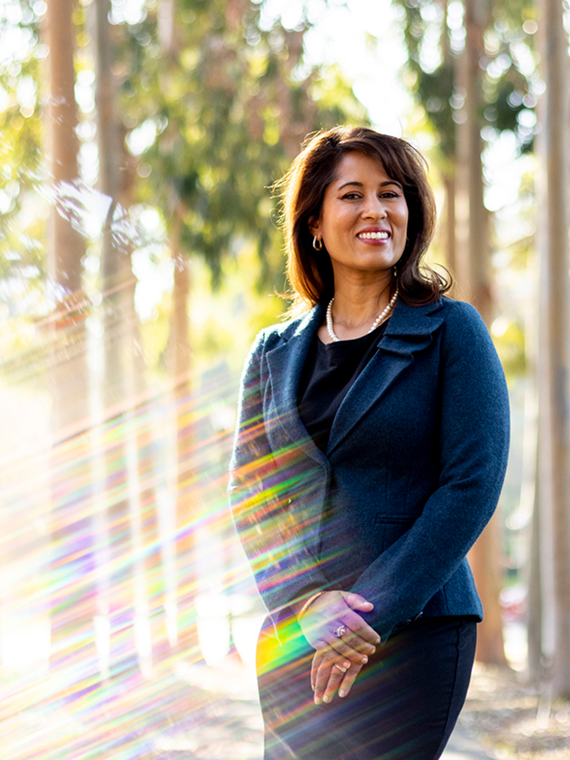 Priya Shimpi wears a dark blazer and dress smiles while standing outdoors among tall trees, with sunlight and colorful lens flares streaming across the image.