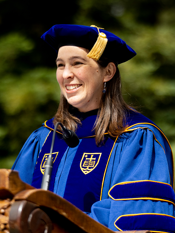 Dean Kochly wears  a blue academic gown and cap with a gold tassel smiles while speaking at a podium during an outdoor graduation ceremony.