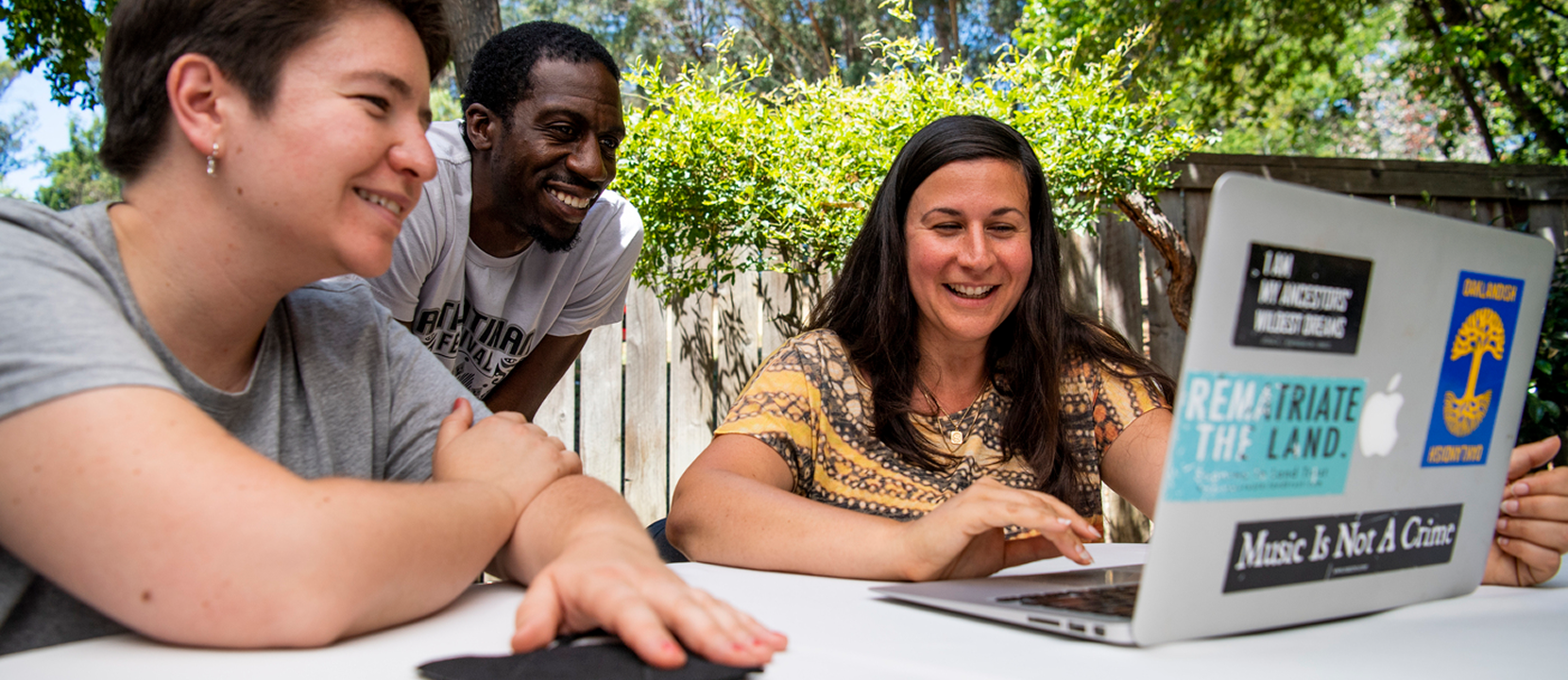 Three people sit and smile around a laptop outdoors at a table, with trees and sunlight in the background. They appear engaged and happy, looking at the computer screen together.