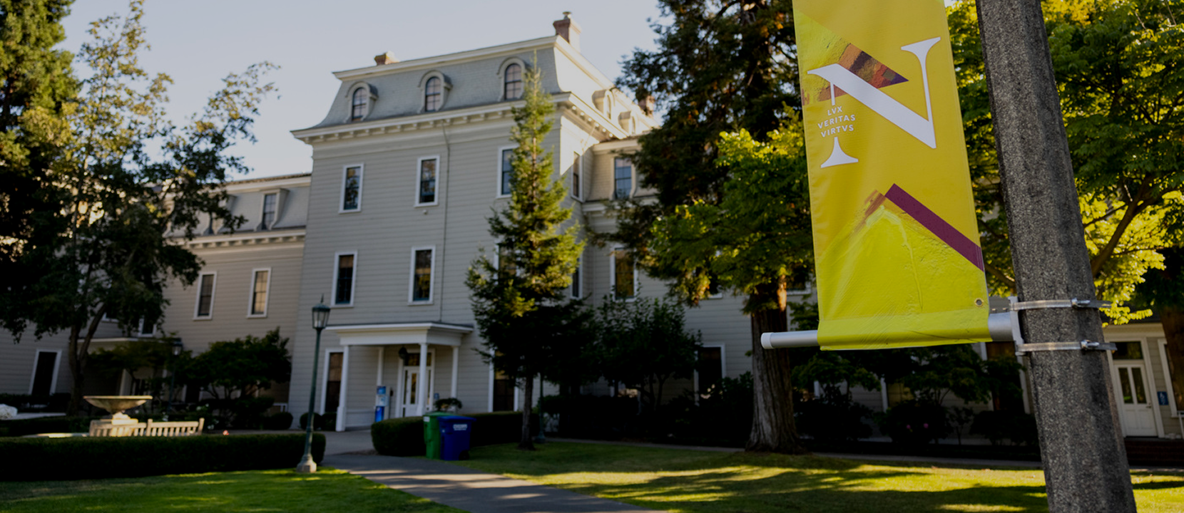 Read view of Mills Hall, a large, historic, light-colored building with many windows is surrounded by trees and a lawn. A yellow campus banner with a large white "N" hangs from a nearby pole in the foreground.