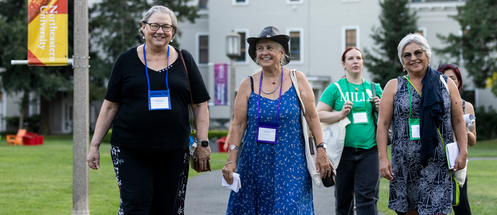 Four women, smiling and wearing conference name tags, walk outdoors on a grassy campus. Other people are visible in the background, and buildings and university banners can be seen.