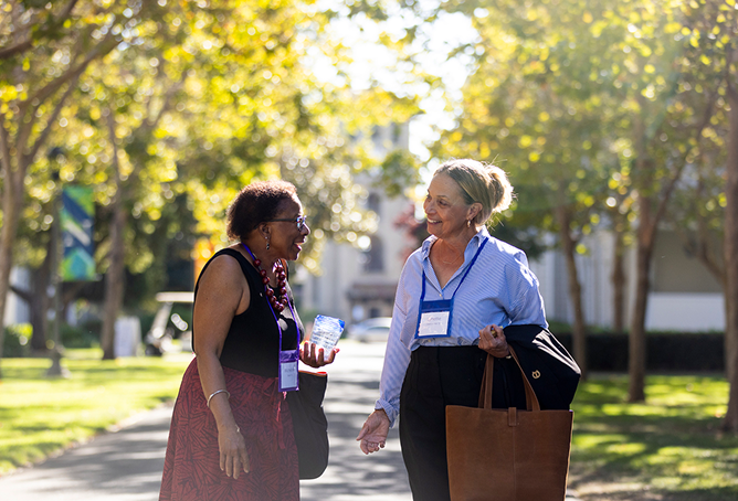 Two women walk and chat on a sunny, tree-lined path. Both wear conference name badges; one holds a drink bottle, the other carries a large brown bag and jacket. They appear to be enjoying a pleasant conversation outdoors.