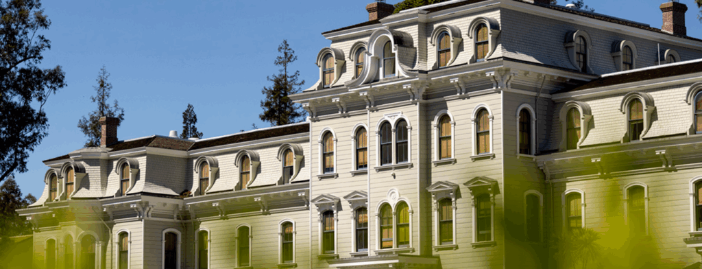 Mills hall, a historic, light-colored building with arched windows, ornate architectural details, and a mansard roof stands under a clear blue sky, partly framed by green foliage in the foreground.