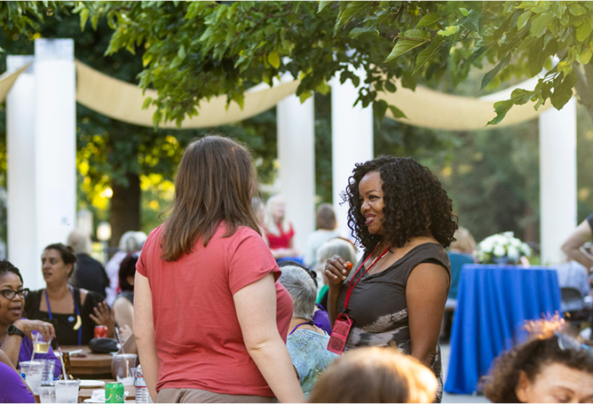 People gather outdoors at an event on a sunny day. Two women are talking and smiling in the foreground, surrounded by others seated at tables under trees and decorative canopies.