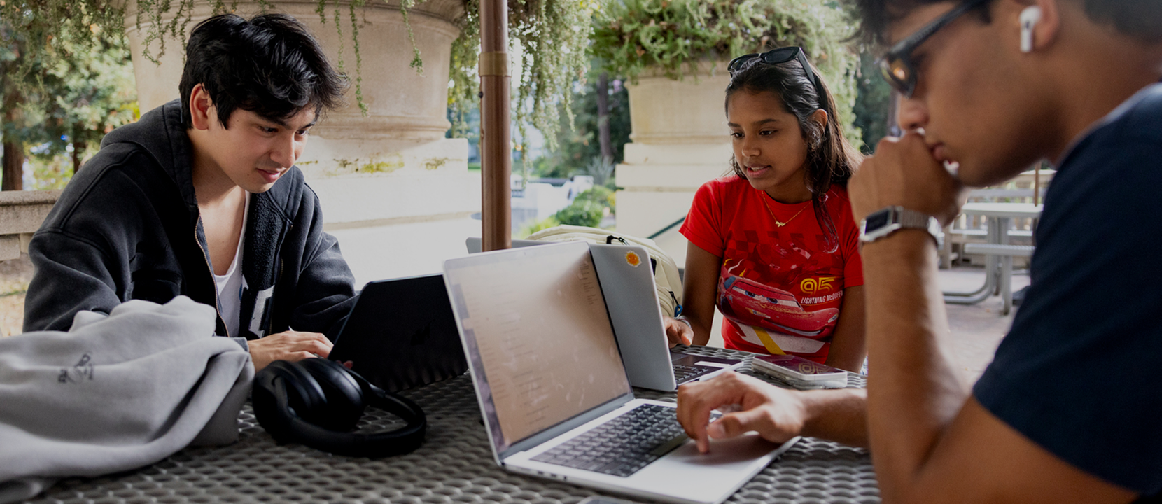 Three students sit at an outdoor table with laptops, studying together. One student wears headphones around his neck, another looks at her laptop, and the third types on his laptop. Trees and greenery are in the background.