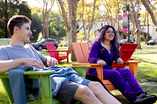 Two people sit outside on colorful Adirondack chairs, talking and smiling. Trees and parked cars are visible in the background on a sunny day.