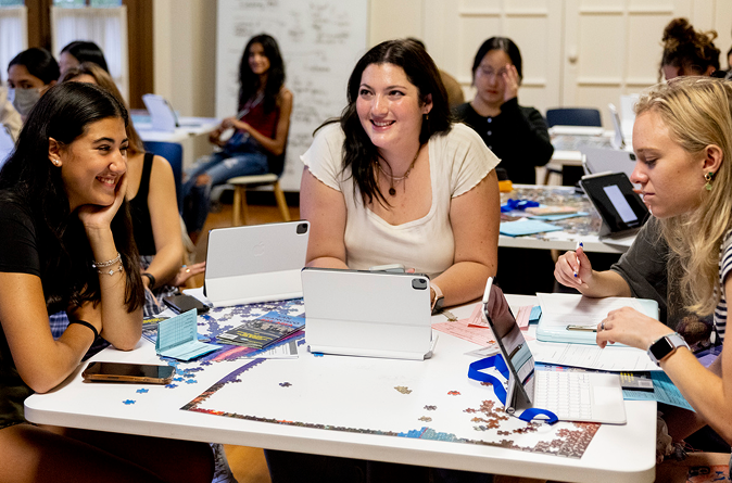 A group of young women sit around a table with tablets, papers, and a puzzle, smiling and talking in a bright classroom with others working in the background.