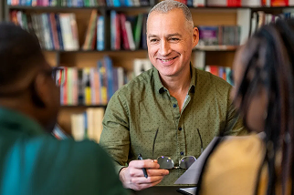 A smiling male teacher holding a book sits at a table with three attentive young students in a classroom setting.