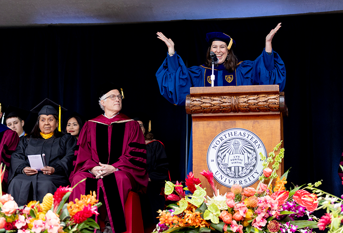Dean Beth Kochly, wearing academic regalia, speaks enthusiastically at a podium with a Northeastern University seal, surrounded by colorful flowers, while Dean Sachs and other people in graduation robes sit beside her on stage.