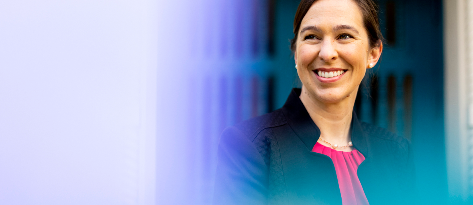 Dean Beth Kochly, wearing a dark blazer and pink blouse, smiles while looking slightly to the side. The background is softly blurred with blue and purple hues.
