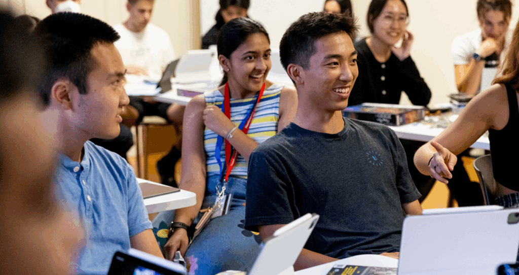 A group of students sit together in a classroom, smiling and engaged in conversation. Several students have laptops open, and one student is pointing while others listen and laugh.