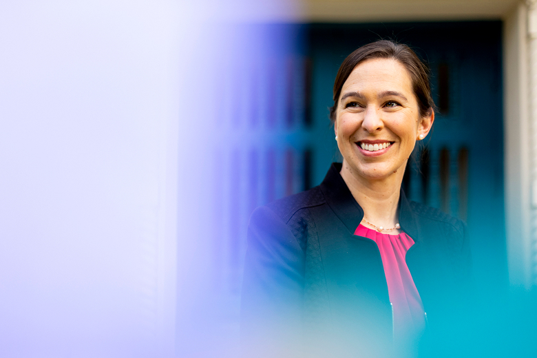 Dean Beth Kochly, wearing a dark jacket, smiles while looking to her left. The background is blurred with a blue door and a soft violet gradient on the left side of the image.
