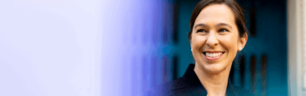 Dean Beth Kochly, wearing a dark jacket, smiles while looking to her left. The background is blurred with a blue door and a soft violet gradient on the left side of the image.