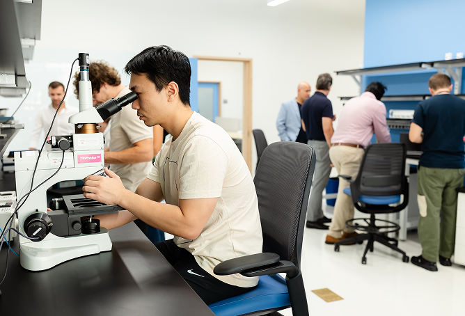 A person sits at a lab table looking into a microscope, while several other people work and talk together in the background of a bright, modern laboratory.