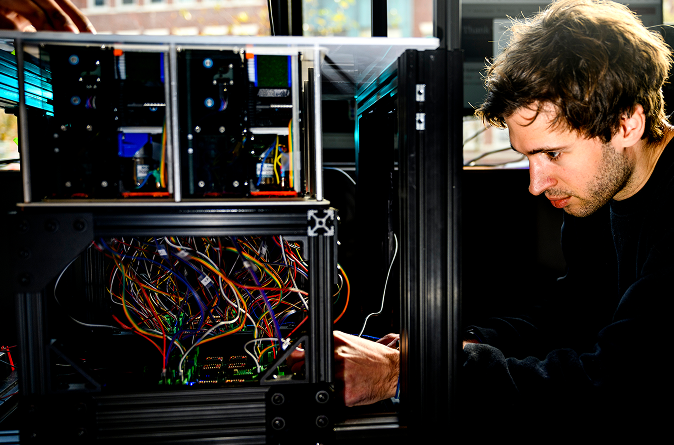 A person intently works with colorful wires and electronic components inside a metal-framed machine, focusing on assembling or repairing complex hardware.