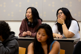 Two women sit at a classroom desk, one wearing headphones around her neck and the other resting her chin on her hand, both attentively listening. Other students are seated in the foreground.