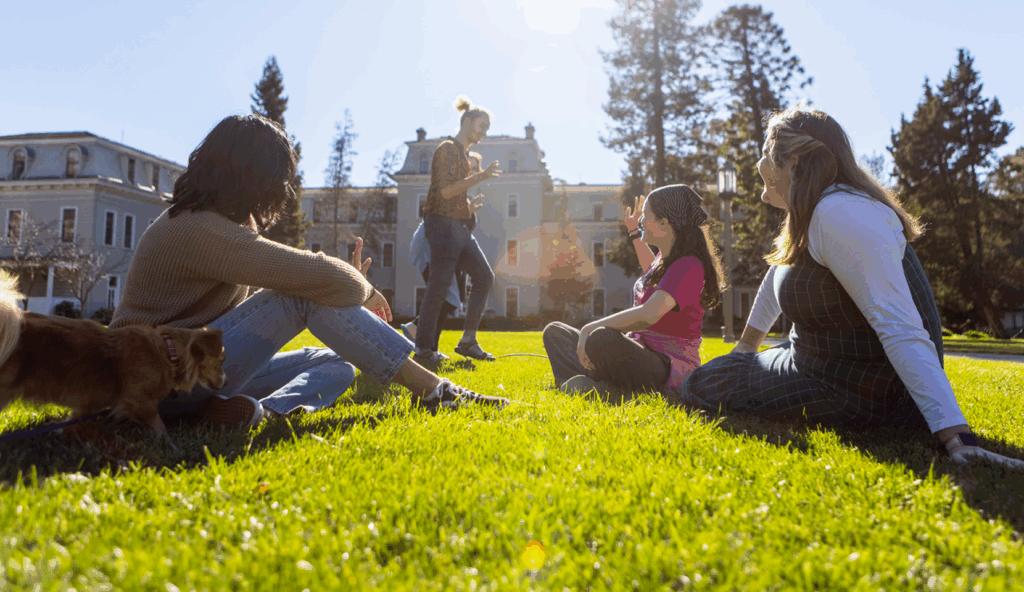 Four people sit and chat on a sunny lawn in front of Mills Hall, with a small dog nearby. Two others stand in the background. Trees and bright sunlight complete the scene.