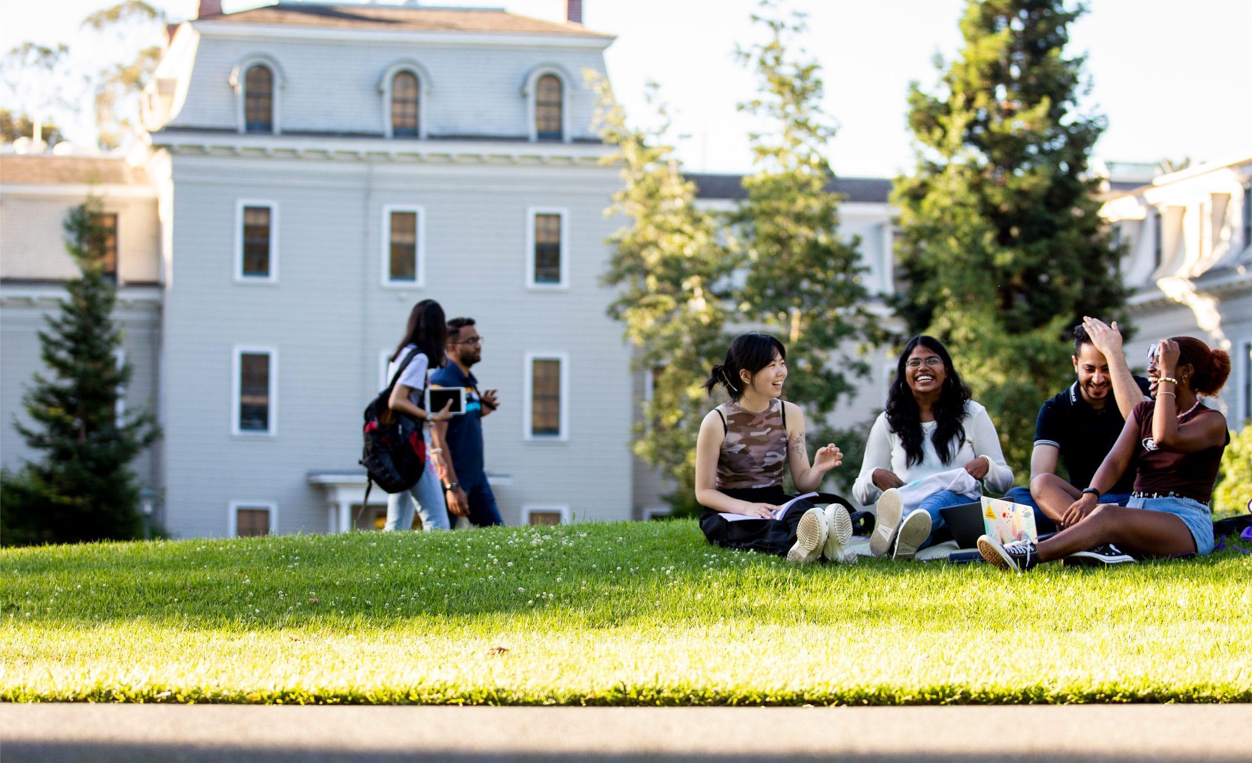 Group of students sits on Holmgren Meadow with Mills Hall in background on Northeastern University's Oakland campus.