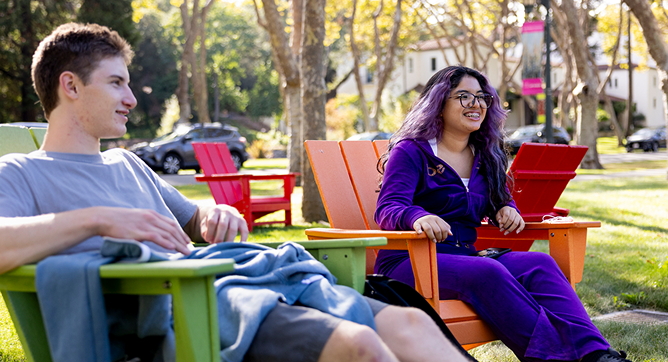 Two people sit outside on colorful Adirondack chairs, talking and smiling. Trees and parked cars are visible in the background on a sunny day.
