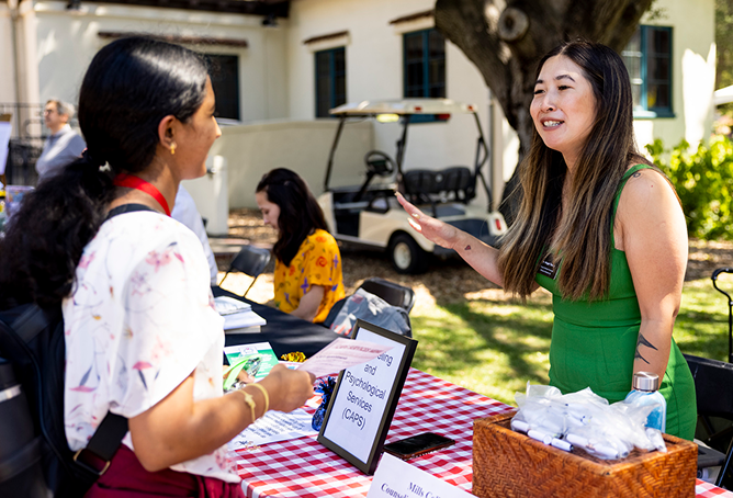 A woman in a green dress smiles and talks to another woman at an outdoor information booth with a red-checkered tablecloth and brochures, under a large tree on a sunny day.