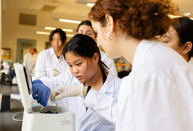 A group of students wearing lab coats and gloves work together on an experiment, focused on a centrifuge in a bright laboratory.