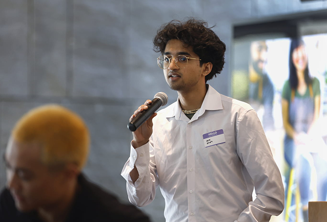A person wearing glasses and a white shirt speaks into a microphone at an indoor event. They have a name tag and appear engaged. Another person with blond hair is blurred in the foreground.