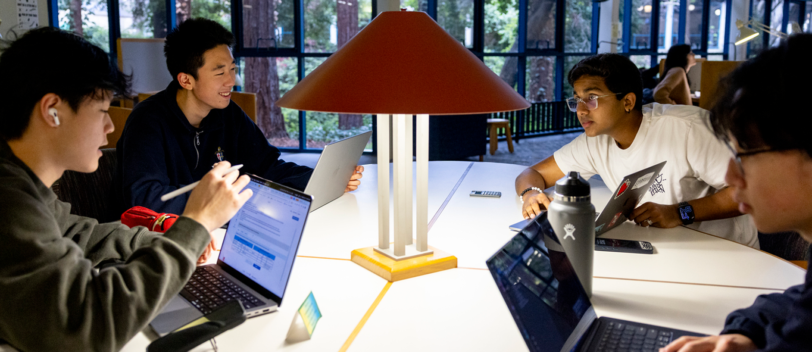Four students sit around a table with laptops and notebooks, collaborating under a large red lamp in a bright, modern study area with large windows and trees visible outside.