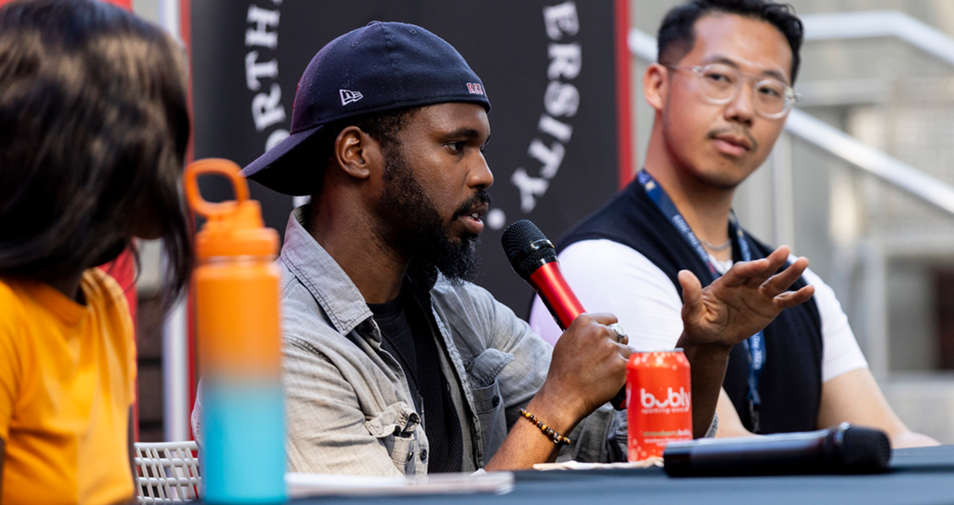 A man speaks into a microphone while gesturing with his hand during a panel discussion, seated between two other people. A bright orange water bottle and a red soda can are on the table in front of him.