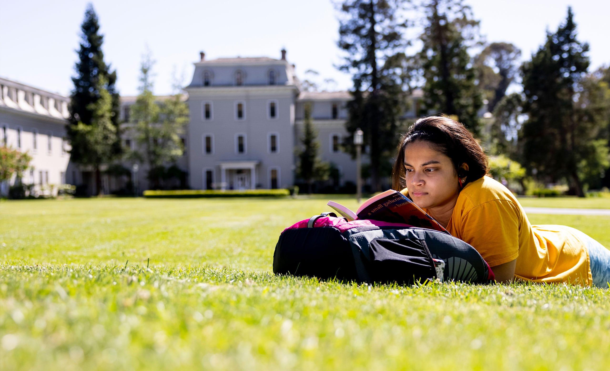 Student lying on the grass of Holmgren Meadow reading with Mills Hall in the distance.