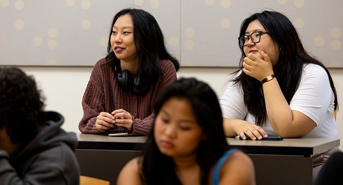 Two women sit at a classroom desk, one wearing headphones around her neck and the other resting her chin on her hand, both attentively listening. Other students are seated in the foreground.